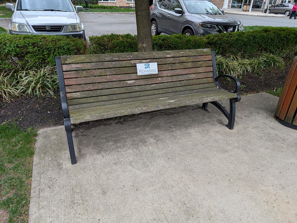 An old wooden bench with moss growing on the wood.  A white placard says "Xchange"