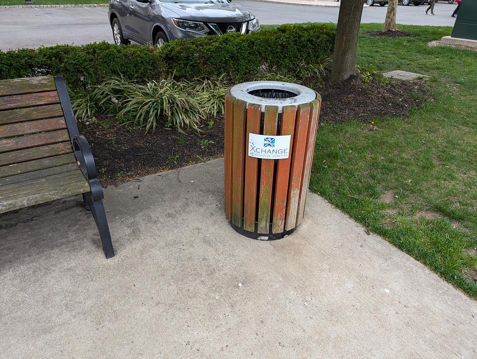 An older wooden trash can, with some moss growing on the wood. The top of the trash can is made of metal.  A black trash bag is visible lining the inside.