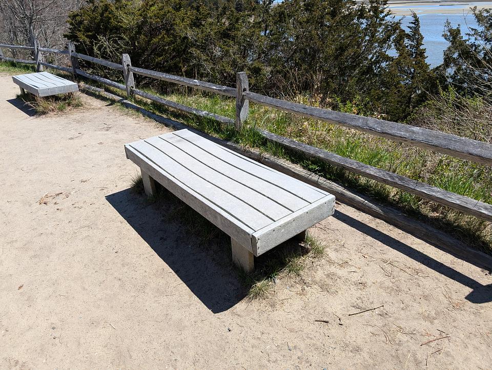 Grey bench made of composite wood in front of a fence, some trees, and the ocean visible far in the distance