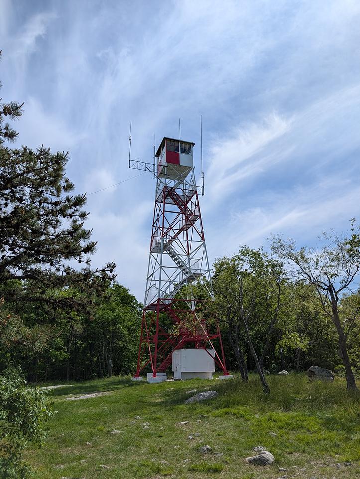 A 4 story fire tower painted in alternating levels of red and white. The top is enclosed with glass windows.  Trees are visible behind the lower levels of the tower.