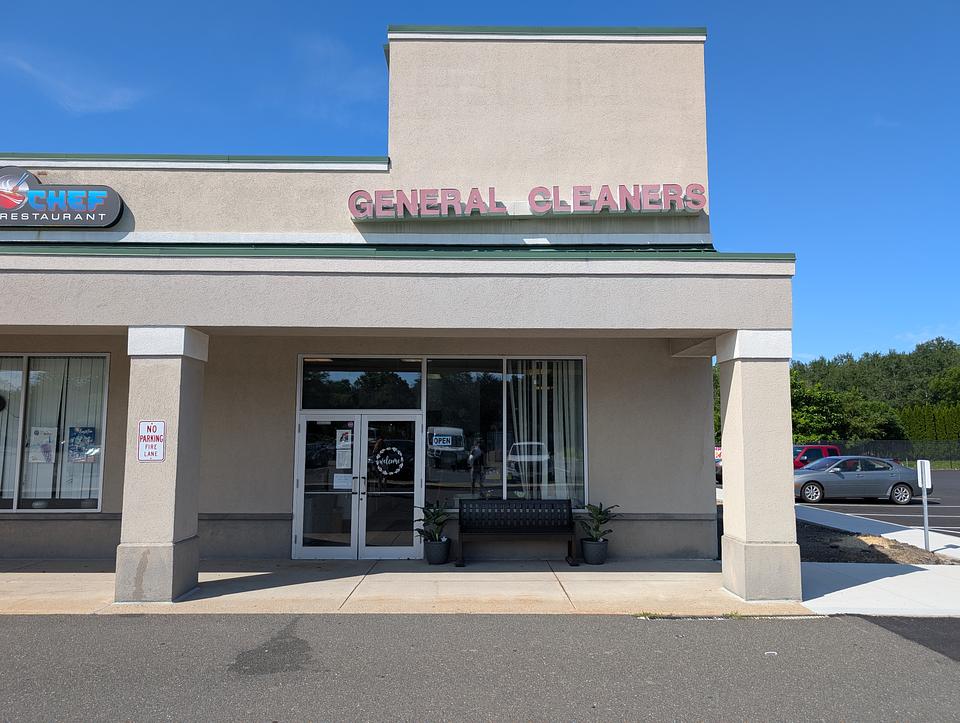 Storefront of General Cleaners.  The name of the business is displayed in large red letters on a grey facade above the windowed storefront. A small amount of green roof can be seen.