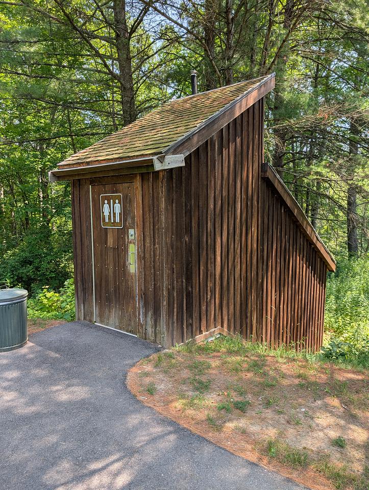 A wooden outhouse with a unisex sign on the door.  The outhouse slopes down an embankment.