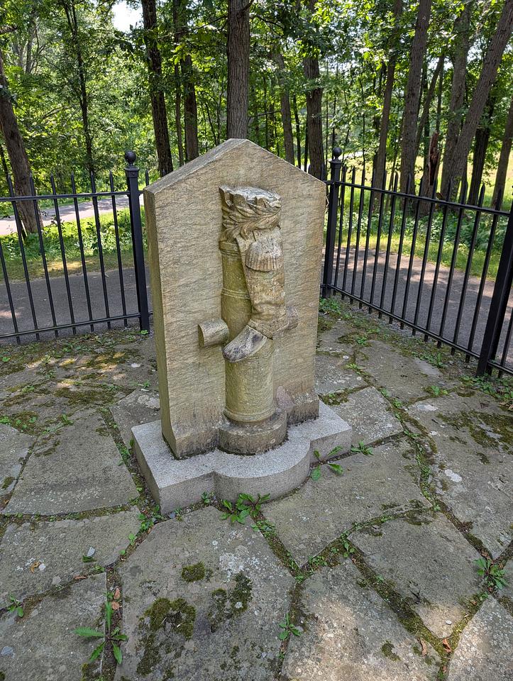 Front view of the boot monument, a white marble stone carved to reveal a riding boot and cannon barrel. The monument is protected by a black metal fence distancing it from a walkway.