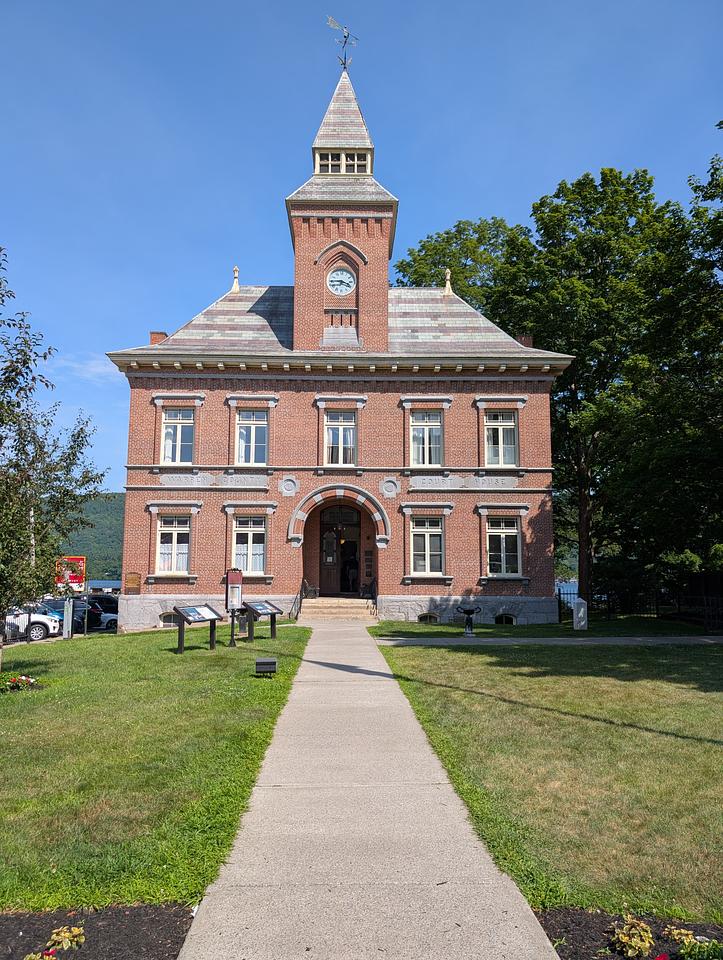 A two-story brick building with a clocktower centered over the top. The clock tower reads 3:45. The first floor of the building has 4 windows with an entryway centered between them.  The second floor has 5 windows visible. A walkway leads up the the building, which historical information signs located along the left of the walkway.