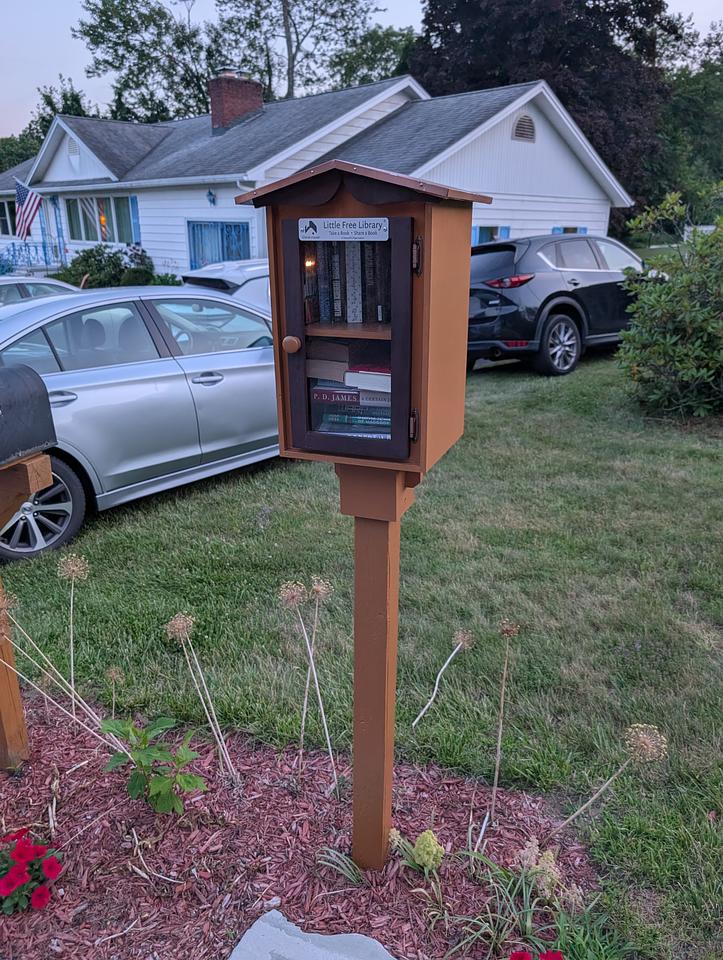 Brown little free library box installed on a wooden post. Several cars are visible in the background.